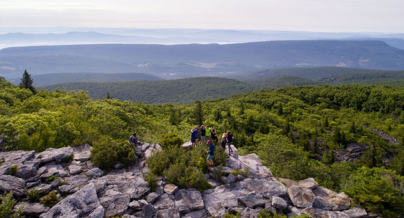 A group of backpackers take in the view of vast trees and mountains from atop a rocky outlook. 